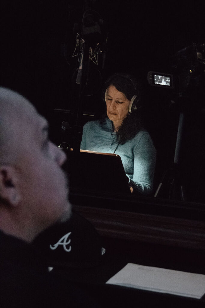 A person in a dark room looks down at a lit screen. They are standing in front of a microphone and are wearing headphones. Another person in the foreground looks on.
