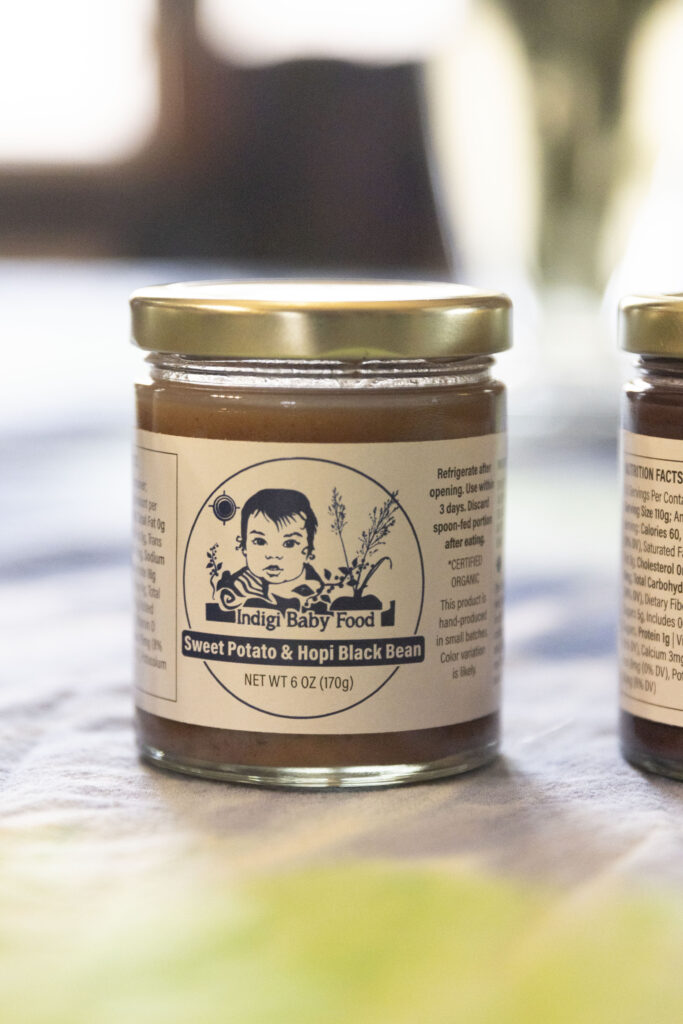 A glass jar containing mushy baby food. The label on the jar has a sketch drawing of a baby and plants. It reads "Indigi Baby Food Sweet Potato and Hopi Black Bean"