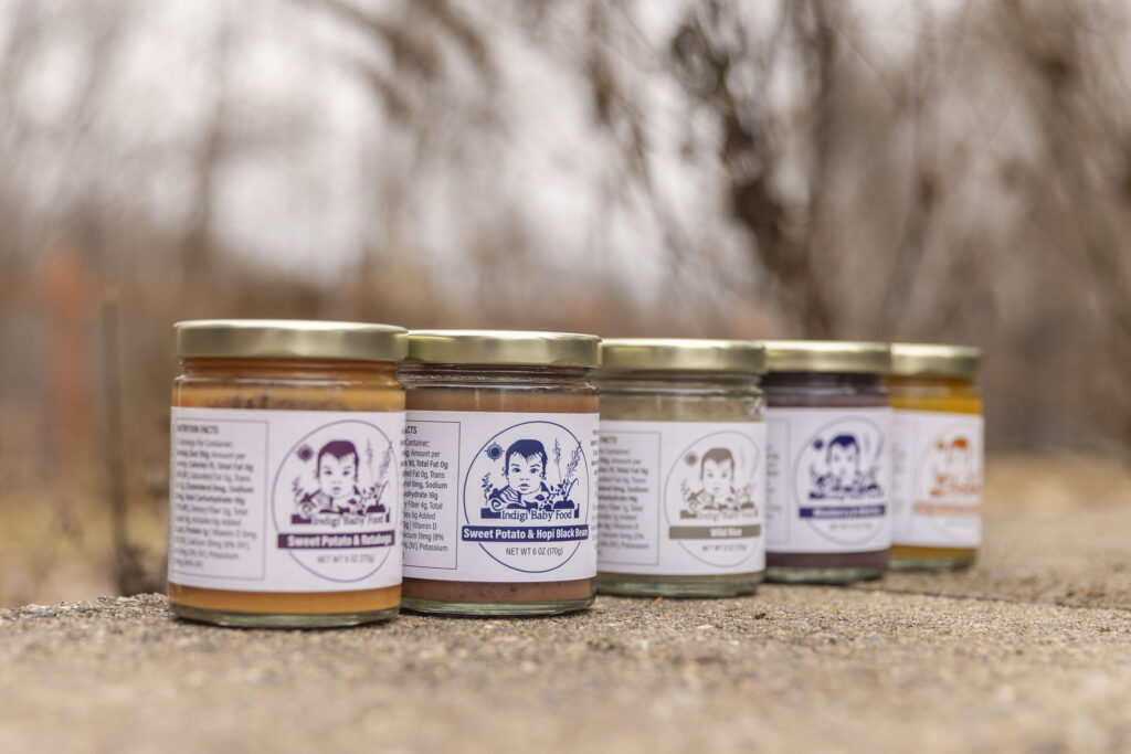 A row of glass jars containing mushy baby food. The contents of the jar for each are different colors, so are the labels. All the labels have a drawing of a baby and plants, and read "Indigi Baby Food" followed by the main ingredients of the food.
