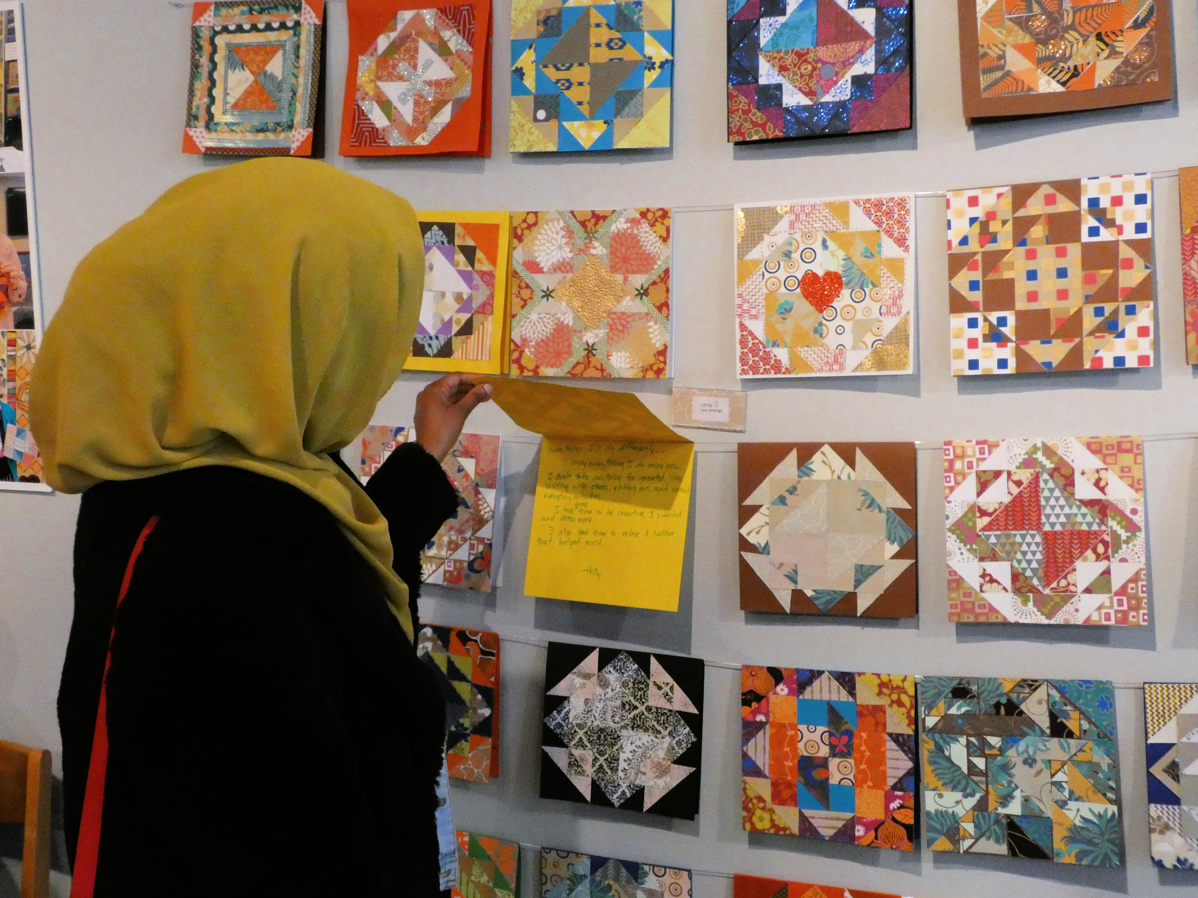 A person wearing a mustard-colored headscarf and black coat faces a wall and interacts with an artwork. The wall has a number of colorful, patterned works of paper, that look like cards, hung on rows of string.