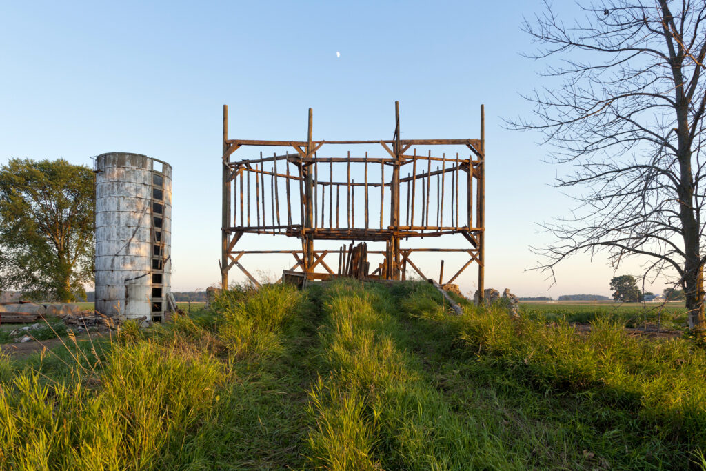 A scaffolding like structure of wooden beams next to a dilapidated concrete grain silo in a farm field.
