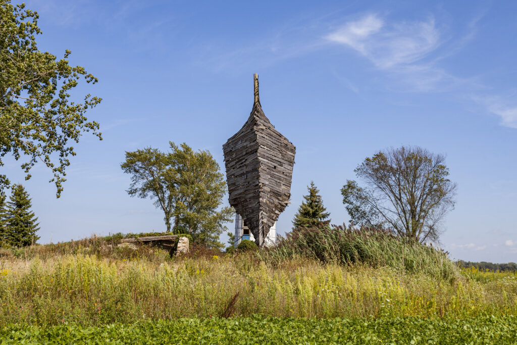 A large structure that looks like an upright ship made of old barn wood stands in a lush overgrown farm field.