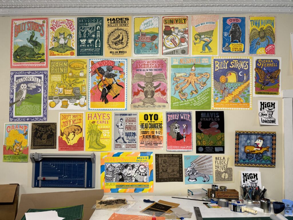 A wall full of letterpress posters. They are colorful, have bold lettering, and some depict animals. There is a table in front of the wall with stationary and other tools.
