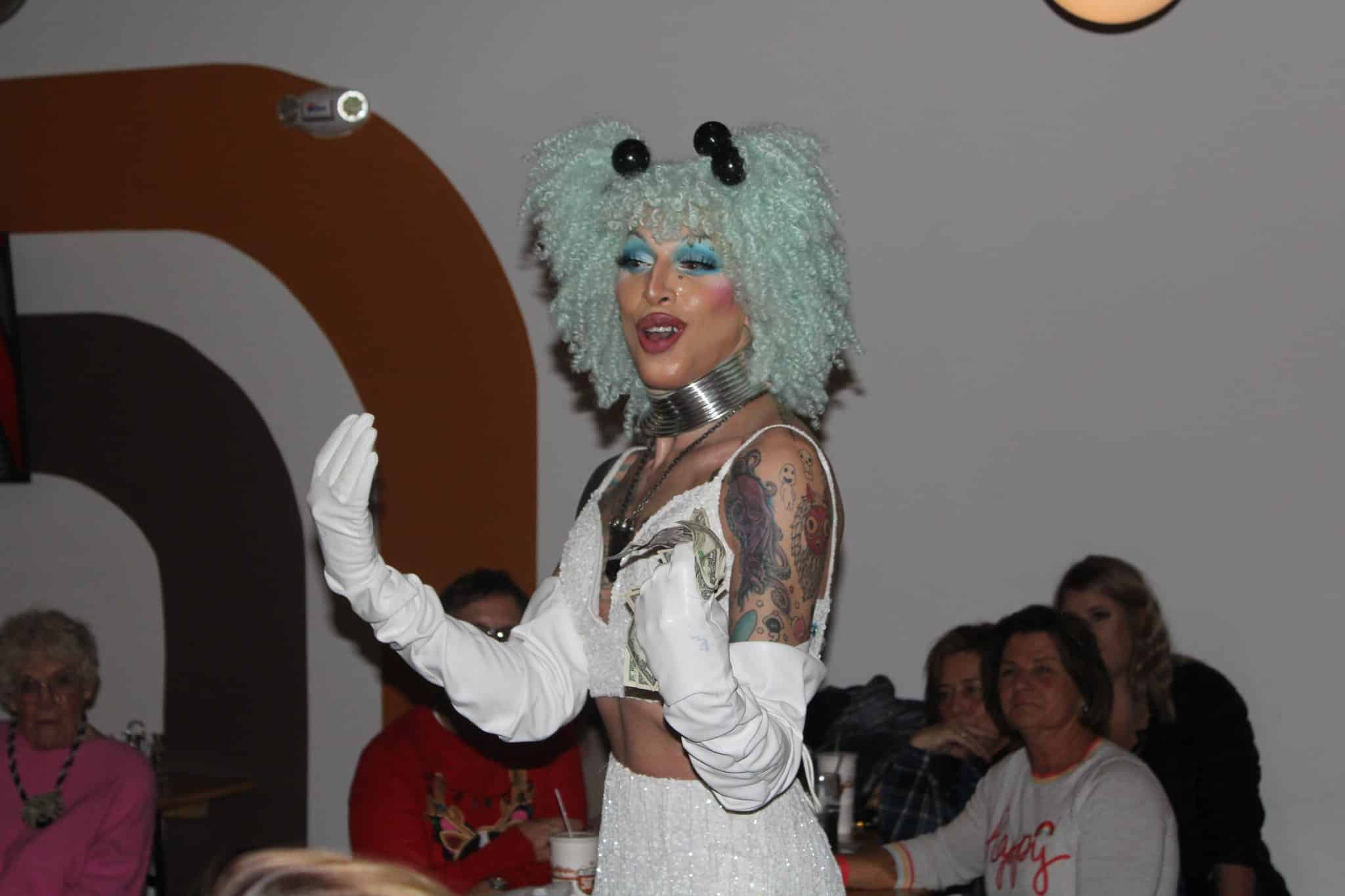 A person in a seafoam green curly wig, white gloves to their elbows, and matching white skirt and crop top stands in the foreground smiling. Several people sit behind them watching.