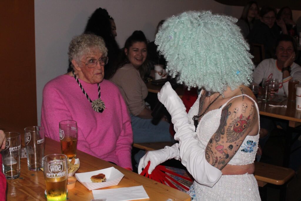 A person in a seafoam green curly wig sits turned towards an older person in a bright pink sweater at a table. Glasses filled with amber liquid litter the table. People behind them smile as they watch them interacting.