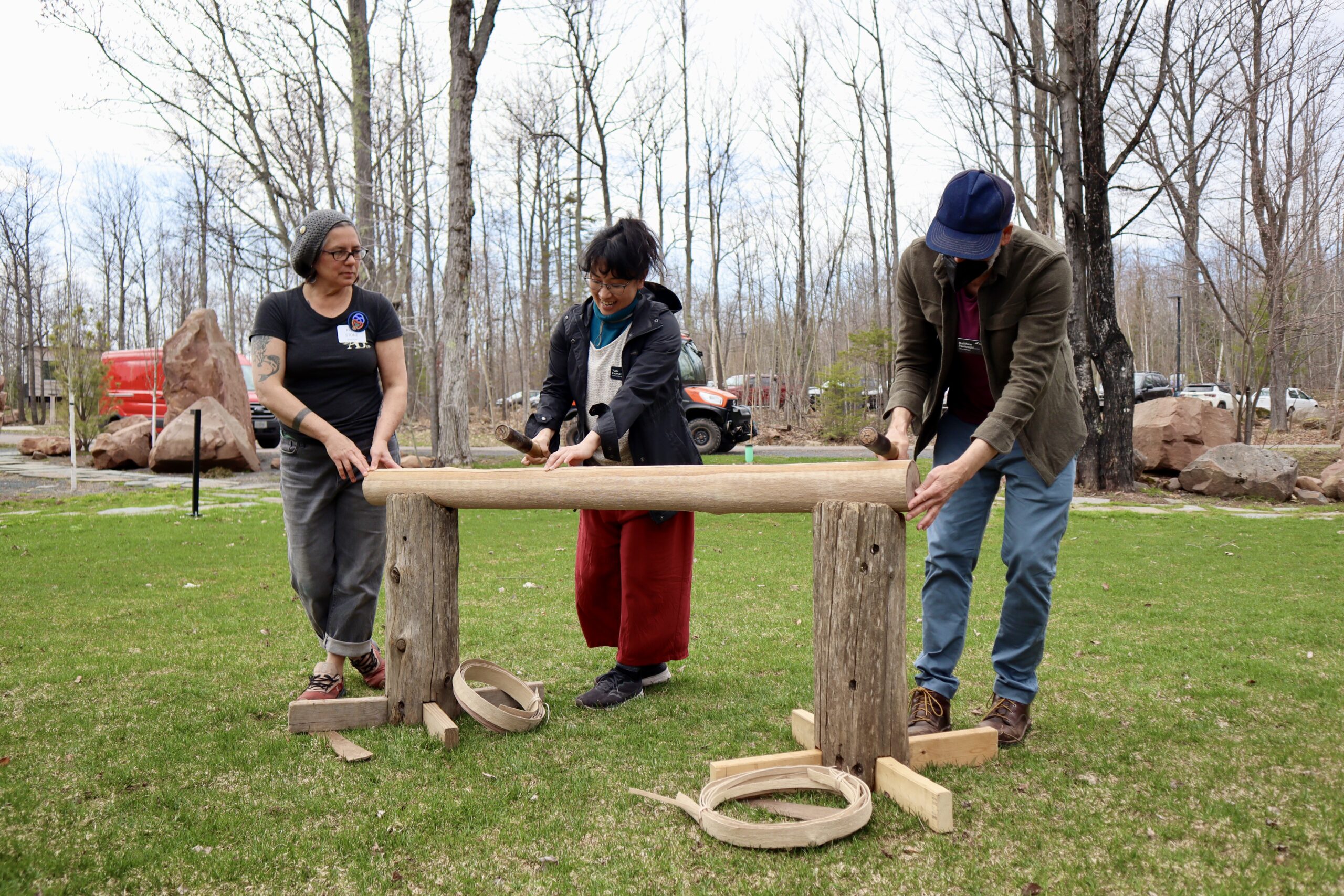 Two people pound a tree to remove a layer of bark for basketmaking as their instructor looks on
