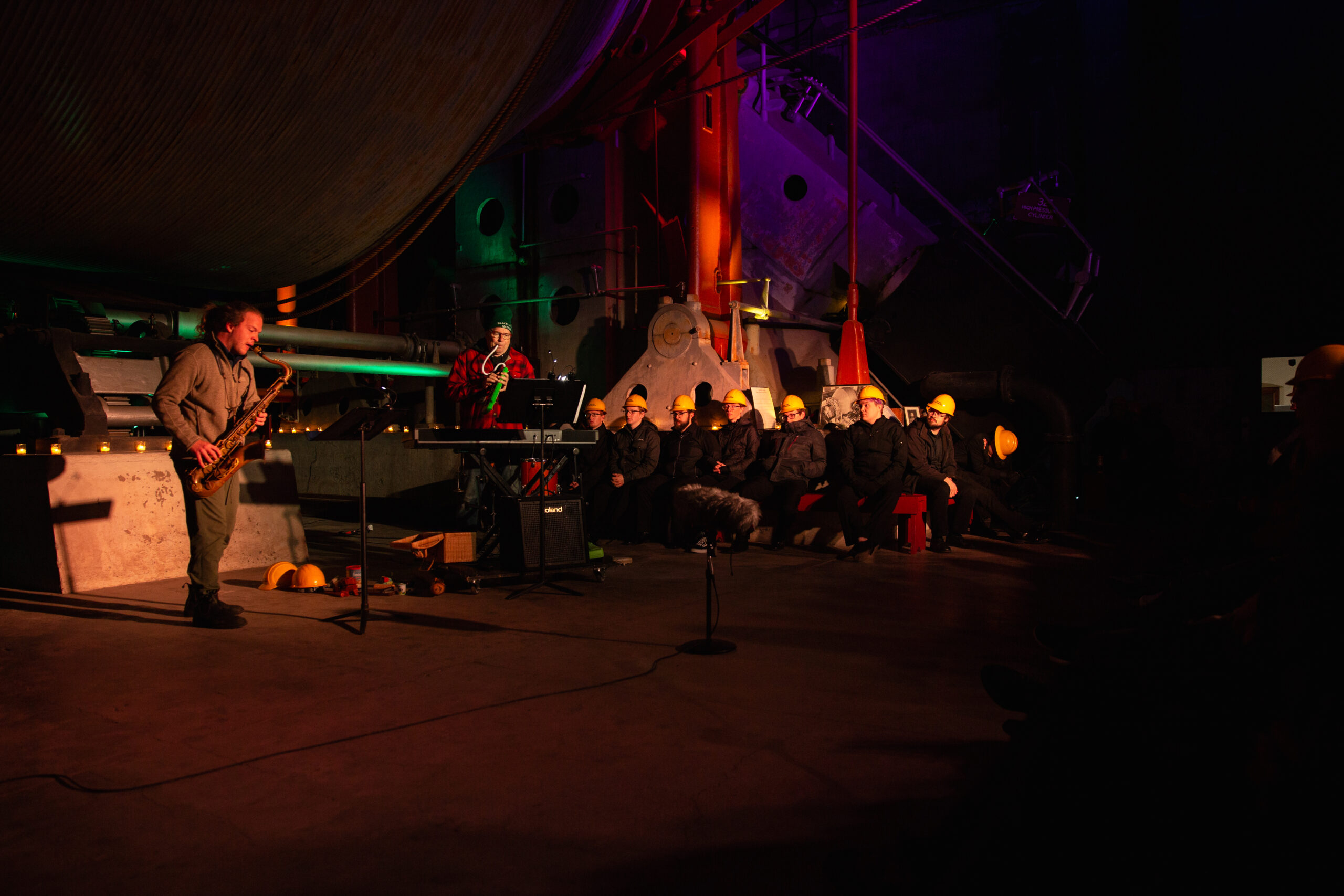 Musicians with wind instruments perform in a dimly-lit space with large industrial equipment. In the background, a group of people dressed in black clothing and yellow hard hats sit on a bench.