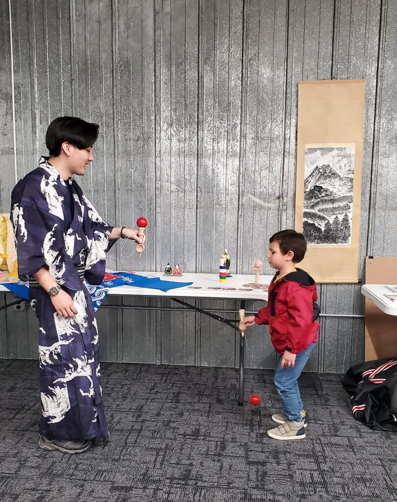 A person of light skin tone dressed in a traditional Japanese robe holds a wooden toy and demonstrates to a child standing in front of them.