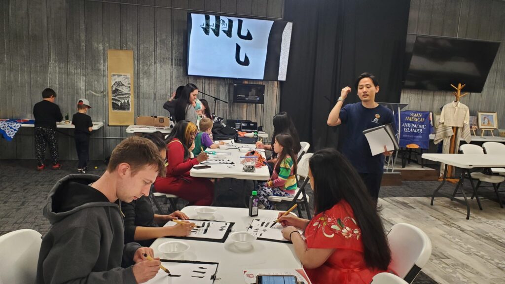 People sit around long white tables and practice writing lettering using paintbrush and black paint.