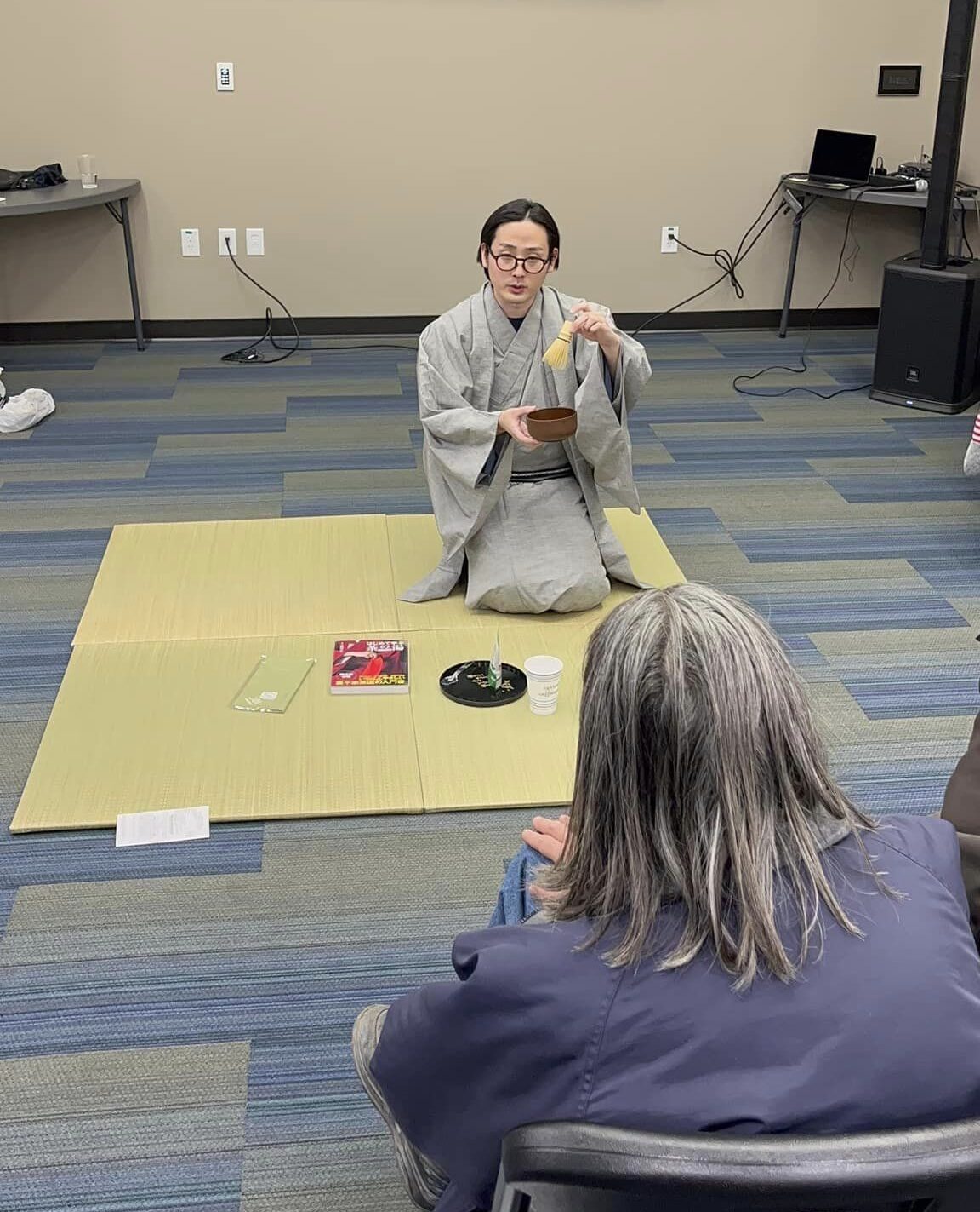 A person of light skin tone wearing a traditional Japanese robe demonstrates the making of tea while sitting on a mat. They are holding a bowl-like vessel in one hand and a small bamboo whisk in the other.