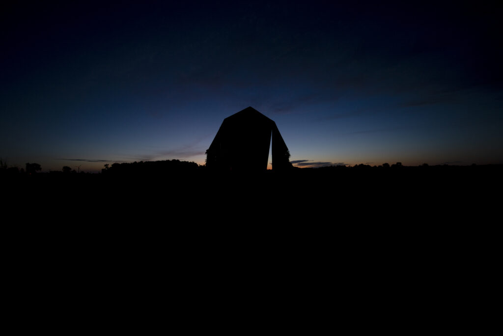 A silhouette of a barn with the sun setting behind it. There is a triangle shaped passage cut into the barn.