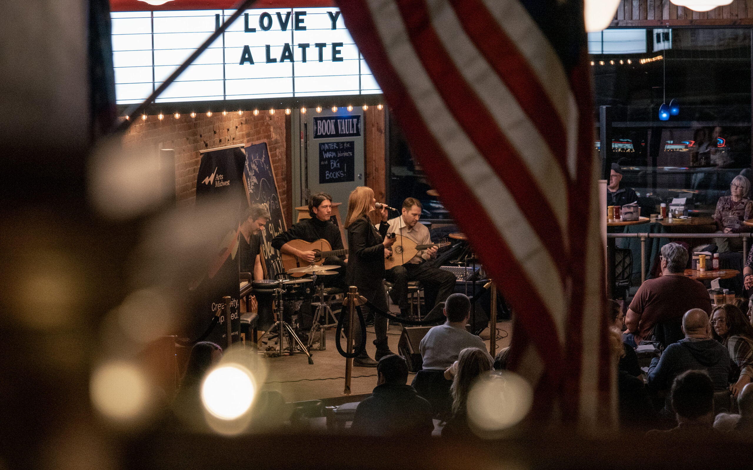 A woman on stage at a coffee shop, shot from a distance with the American Flag in focus