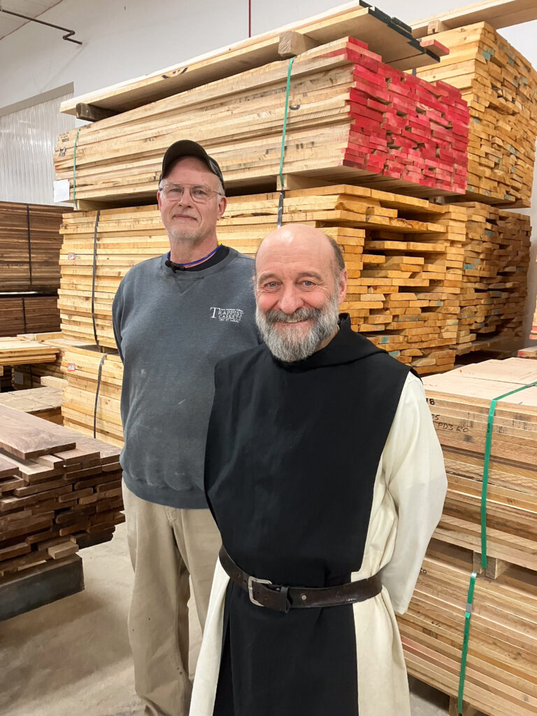 Two people of light skin tone standing and smiling. One is wearing a black and white robe, and the other is in a dark grey sweatshirt and khaki colored pants. In the background are stacks of lumber.