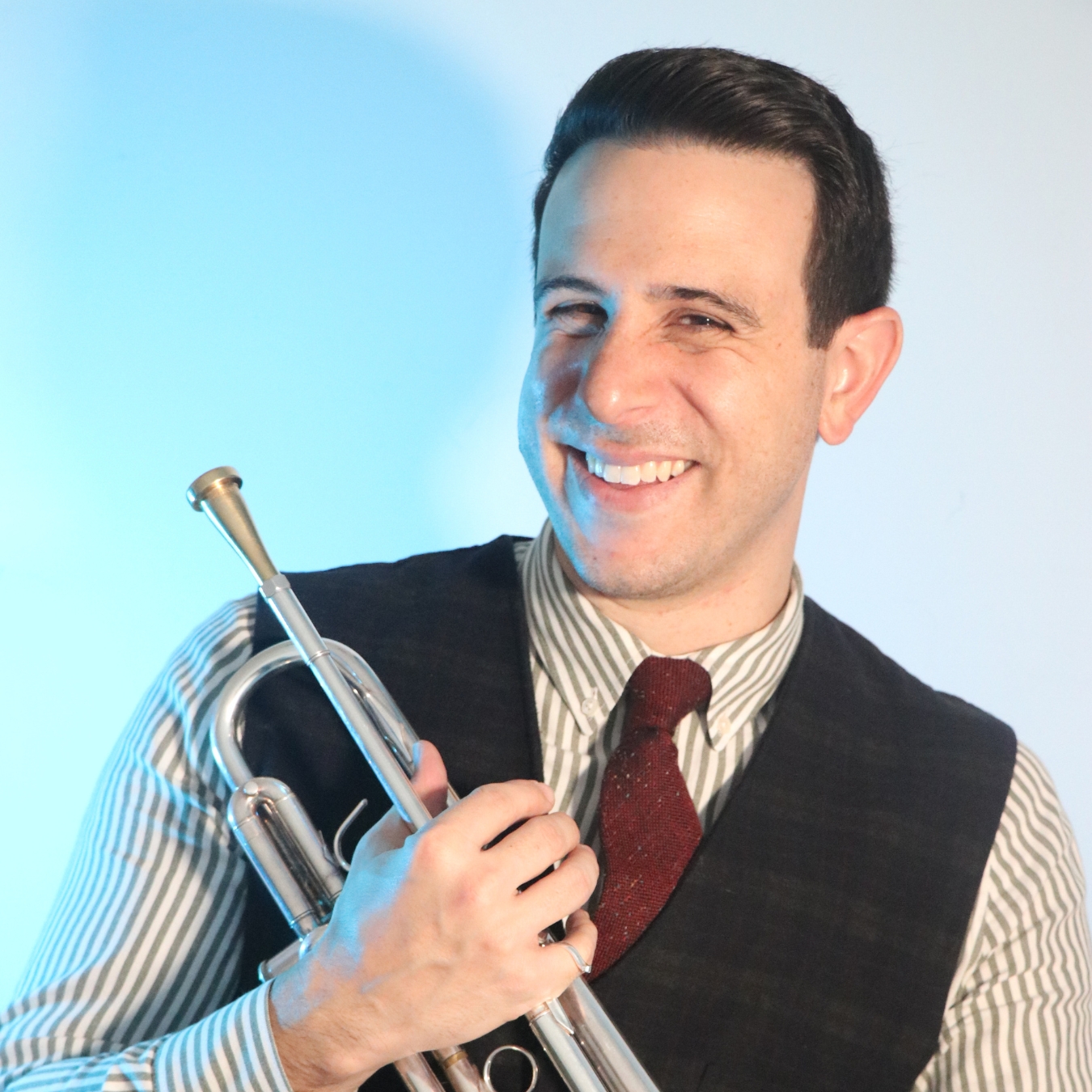 A person of light skin wearing a striped button down, gray sweater vest, and red tie; smiling and holding a trumpet.