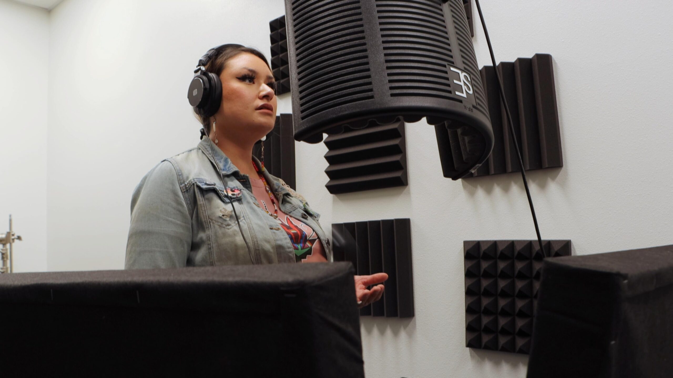 A person in a jean jacket stares at recording equipment in front of their mouth in a sound booth. Their mouth is slightly open and they might be talking or singing.