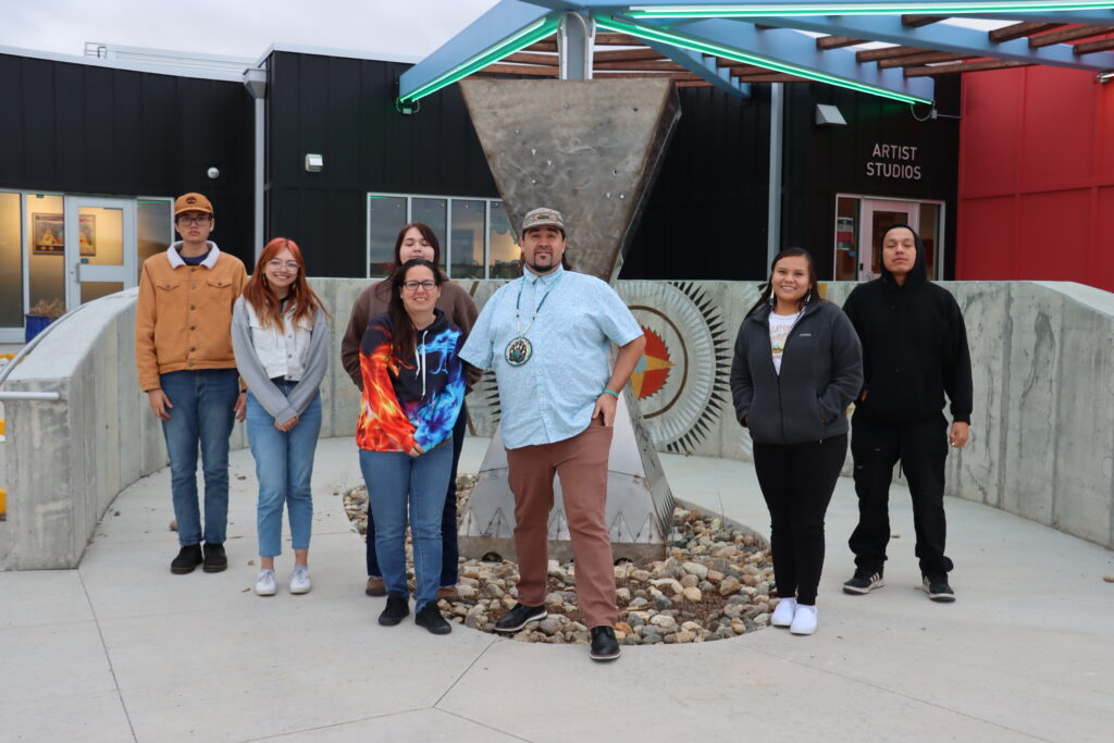 A group of people stand outdoors in front of a building with a circular cement sculpture and hour-glass shaped metal sculpture behind them. The words "Artist Studios" appear above a door to their right.