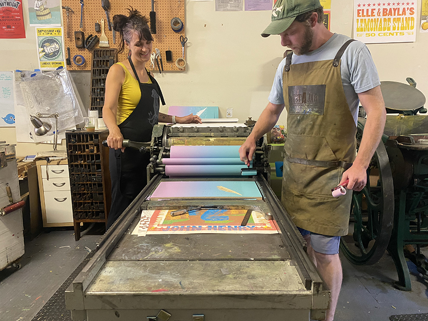 Two people stand across from each other looking at and operating an old letterpress printing table in between them.
