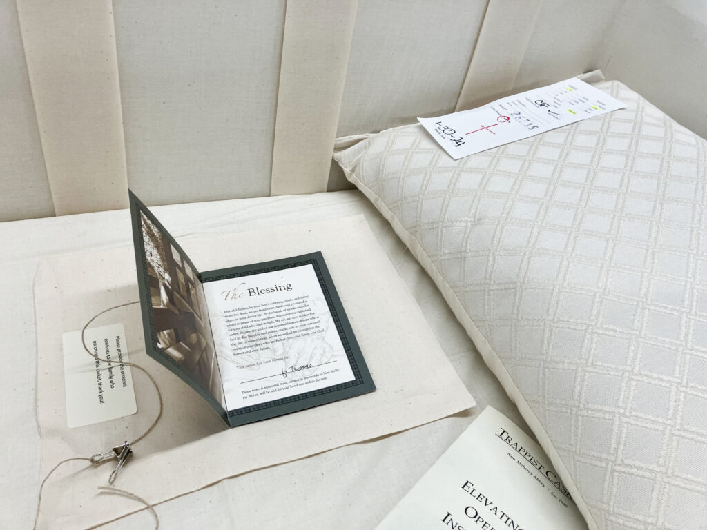 The inside of a casket upholstered with off-white fabric and a pillow with a textured fabric cover. There is a card laid inside with words and a title that reads "the blessing."