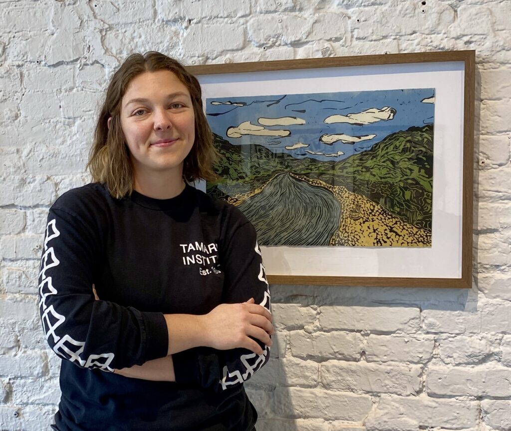 A person in a longsleeve black t-shirt smiles with their mouth closed in front of a white brick wall and a framed print of a river landscape of blue, green, and yellow-gold.