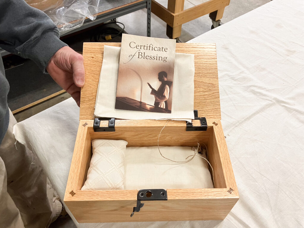 A hand holds open a small wooden box. There is a small white pillow and pad inside the box.