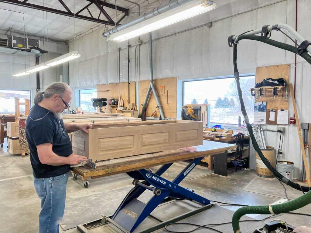 A person of light skin tone wearing a black tshirt and jeans works on building a wooden casket in a woodshop.