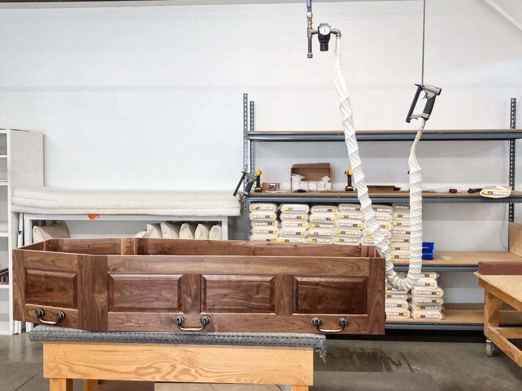 A dark wooden casket, in process of getting built and finished, sits on a table.