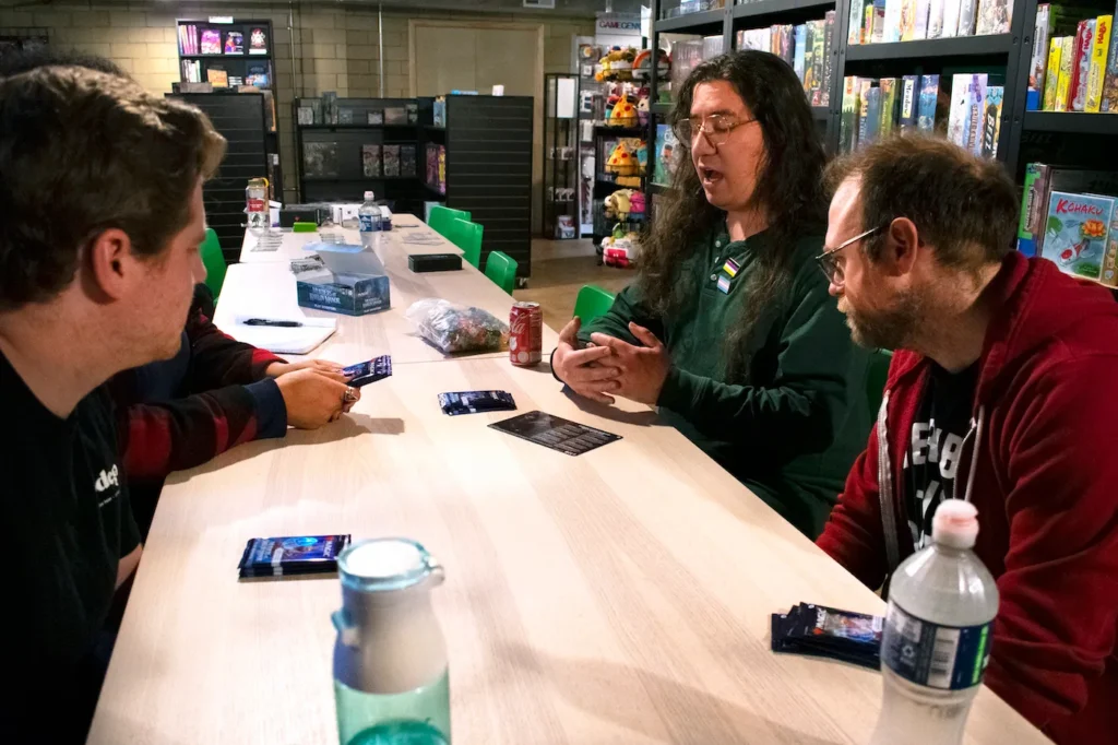 Four people sit gathered around a wooden table with cards and water bottles in front of them. One person is talking and the others are looking at them.