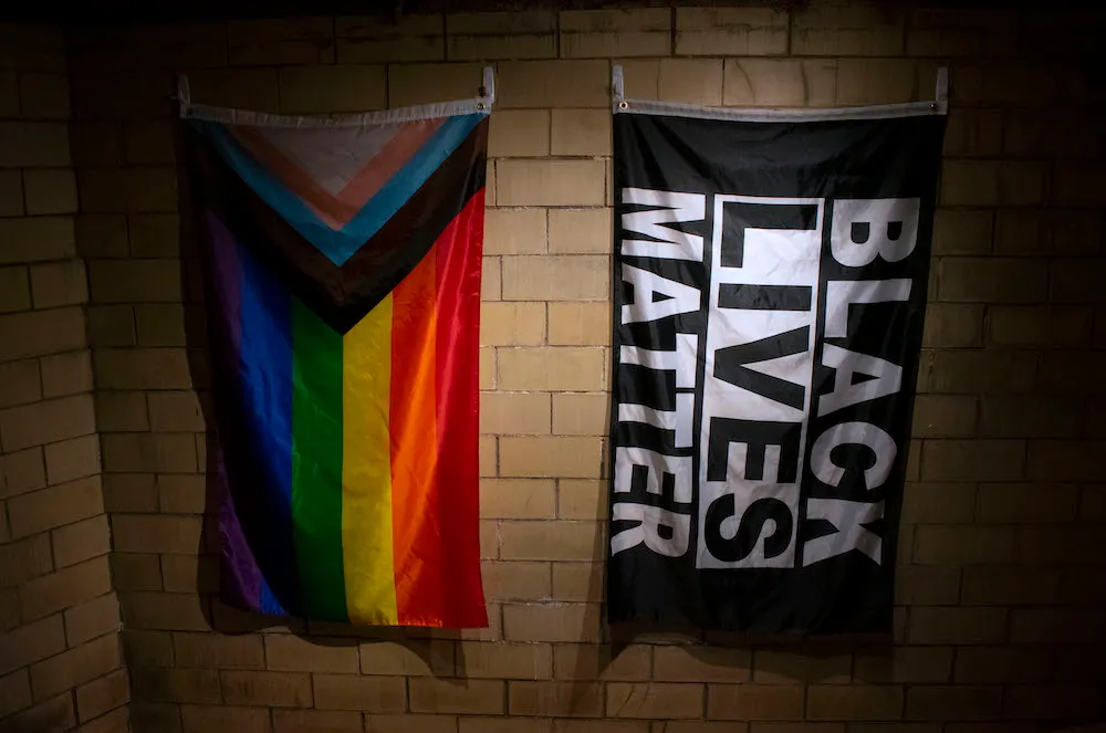 An LGBTQ+ Pride Flag and a Black Lives Matter Flag hang in front of a brick wall.