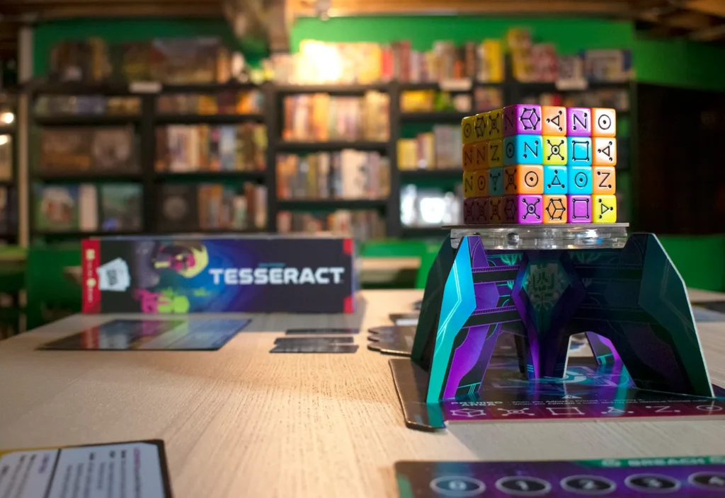 A brightly colored game of Tesseract sits on a wood table. In the background bookshelves are stacked with a wide variety of other board games.