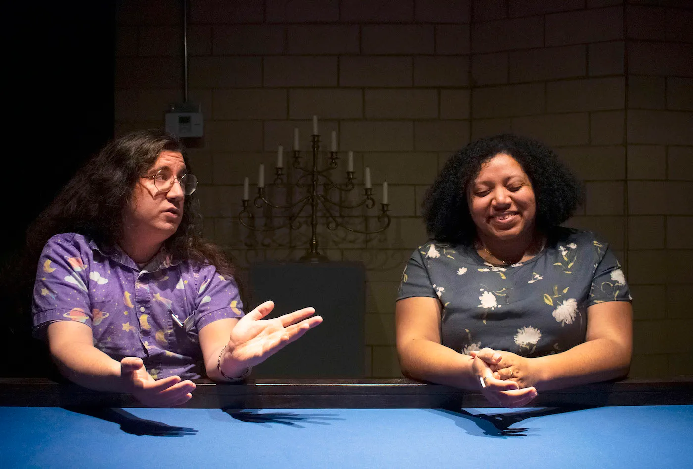 Two people sit in front of a blue table under harsh lighting. One person is raising their arms to explain something, the other has their hands clasped and is smiling.