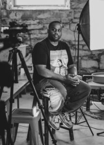 A person with dark skin tone and short, buzzed hair wearing a t shirt and cargo shorts and sitting on a stool in a portrait studio.