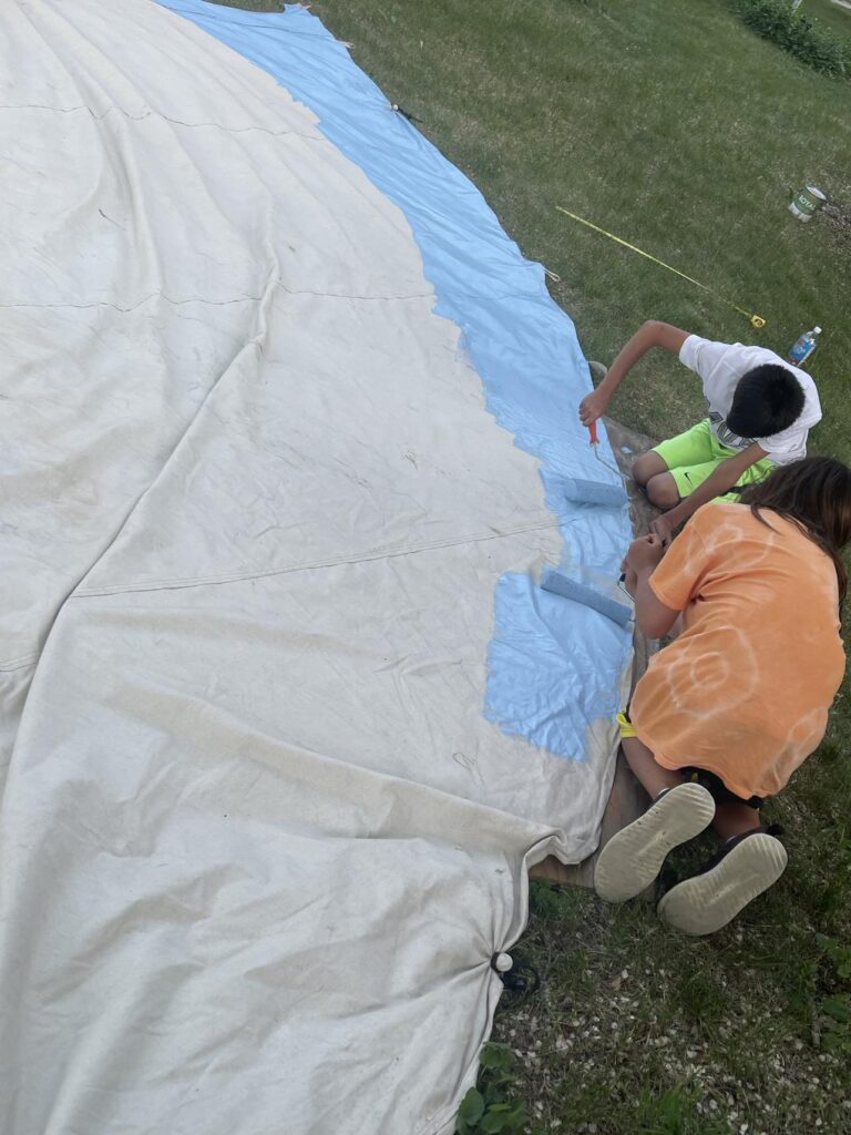 Two young people using paint rollers to roll sky blue paint onto a large piece of fabric.