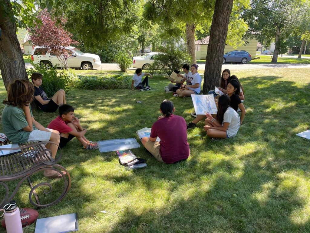 Twelve youth sitting in a circle on the ground in a greenspace, holding drawings.