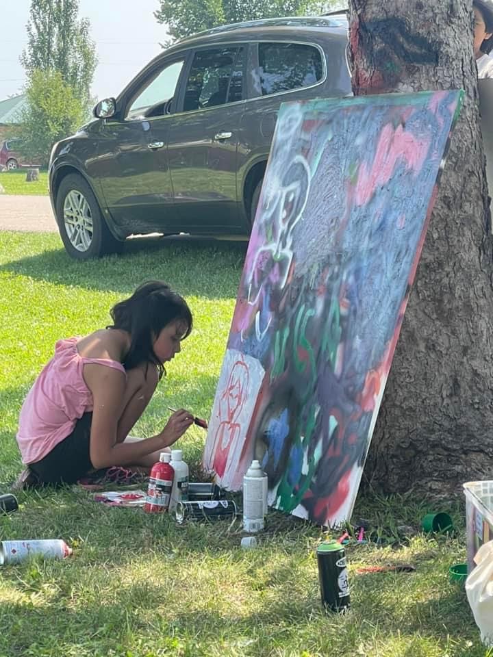 A young girl sitting in the grass, using a paintbrush to outline a figure on a large piece of canvas.