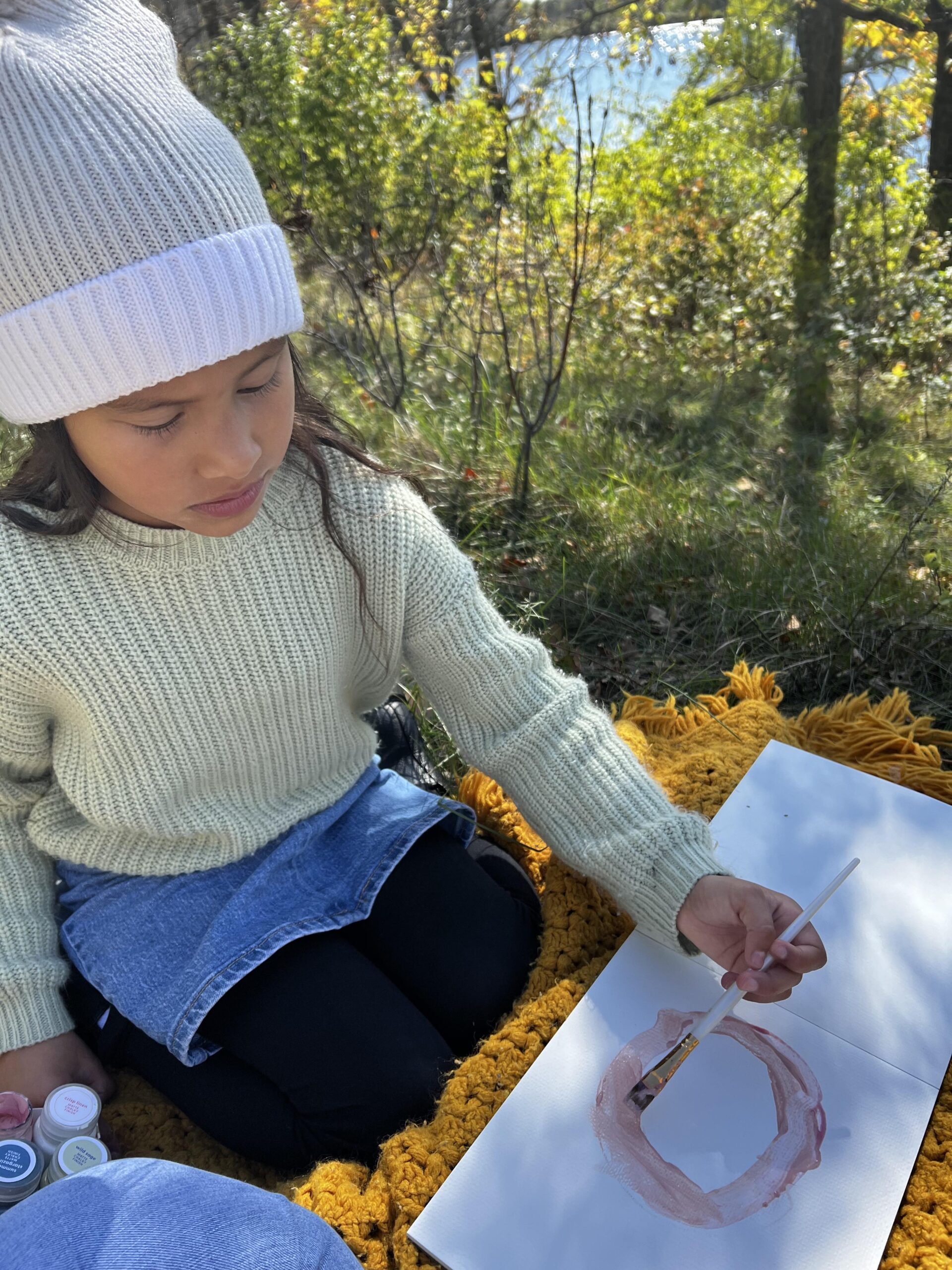 A young girl wearing a sweater, denim skirt, and beanie, painting a circle on a piece of paper.
