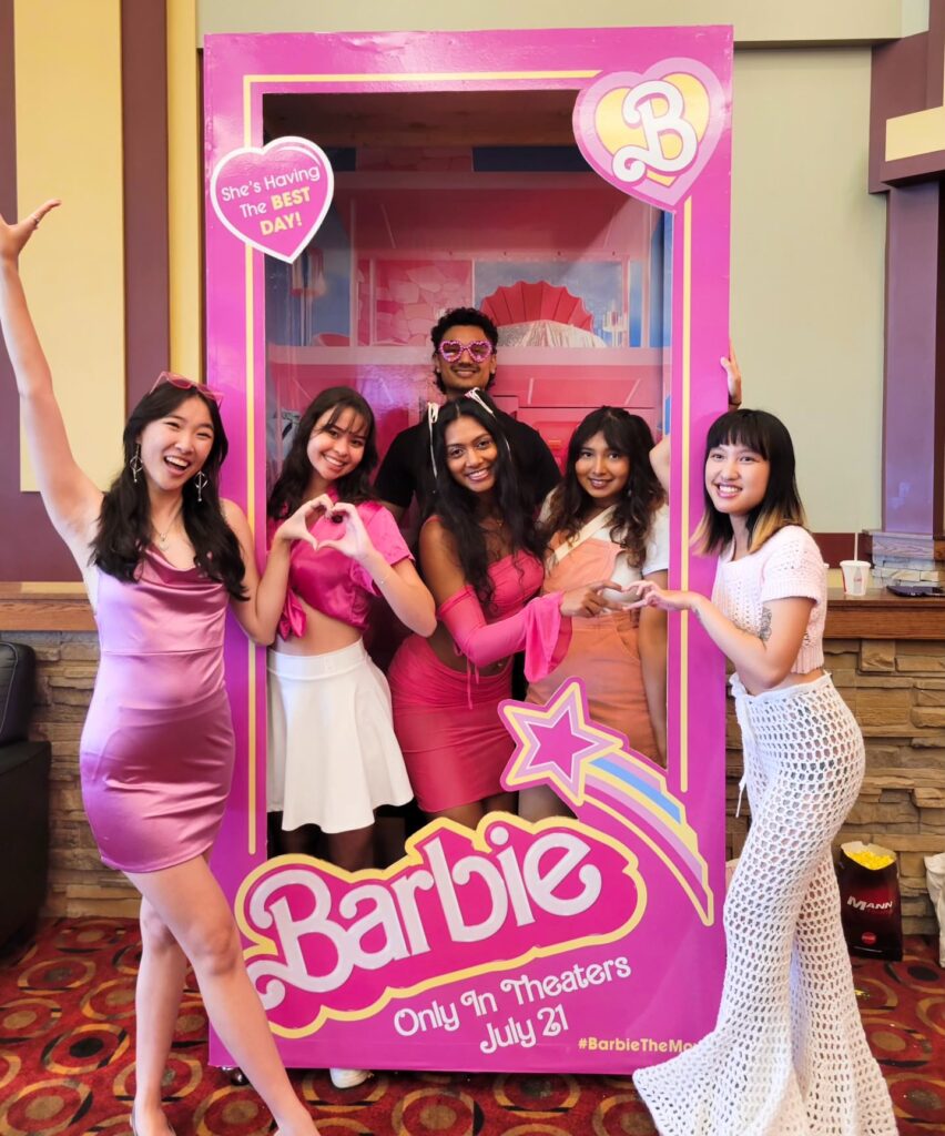 Six people dressed in pink and white outfits standing in and around a promotional standee shaped like a Barbie toy box, with signage reading, "Barbie: only in theaters July 21."
