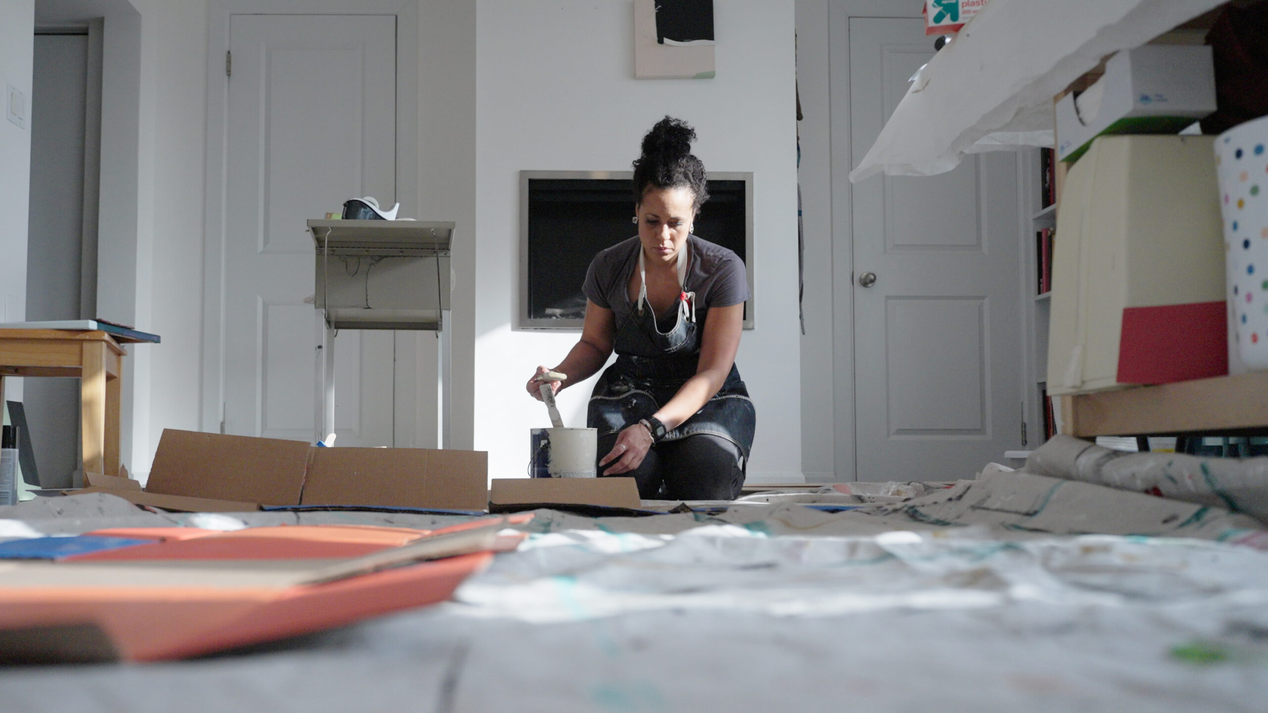 A person kneels before a large canvas holding a thick wide brush caked in white paint. They are wearing a dark jean apron. Sun dapples the canvas, the contents of which are not visible to the camera.