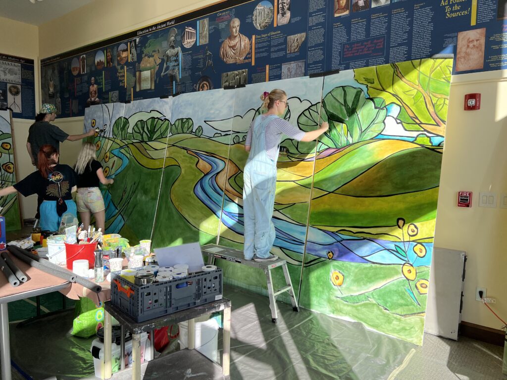 A person paints trees on a mural panel