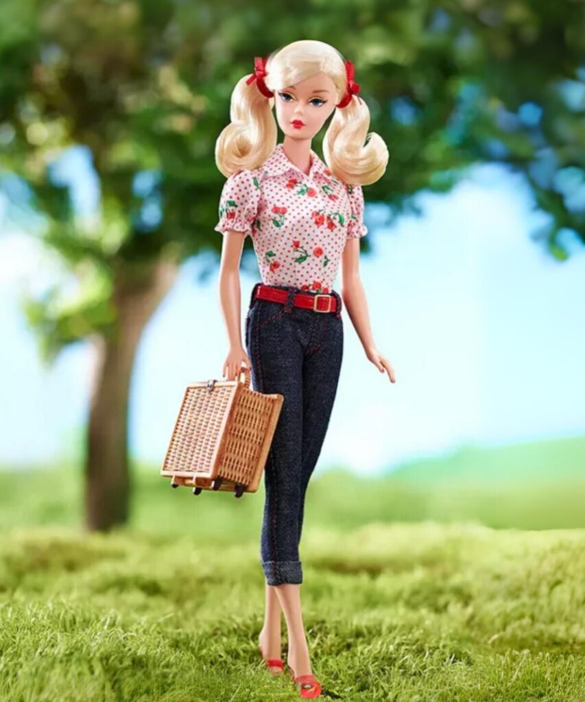A Barbie doll with pigtails wearing a cherry-pattened blouse and jeans, carrying a picnic basket.