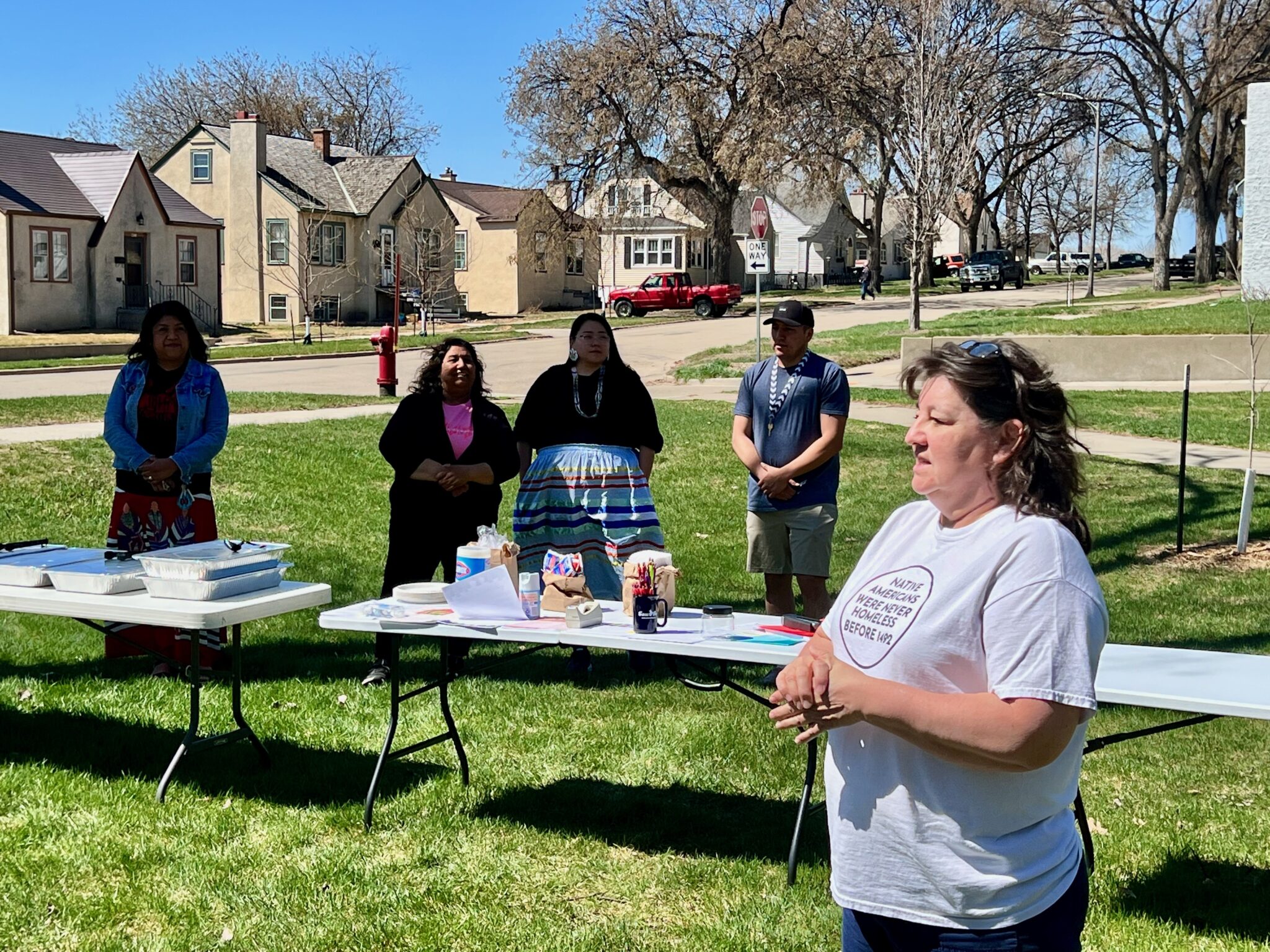A woman of medium light skin tone speaks at an outdoor meeting filled with folding tables and community members.
