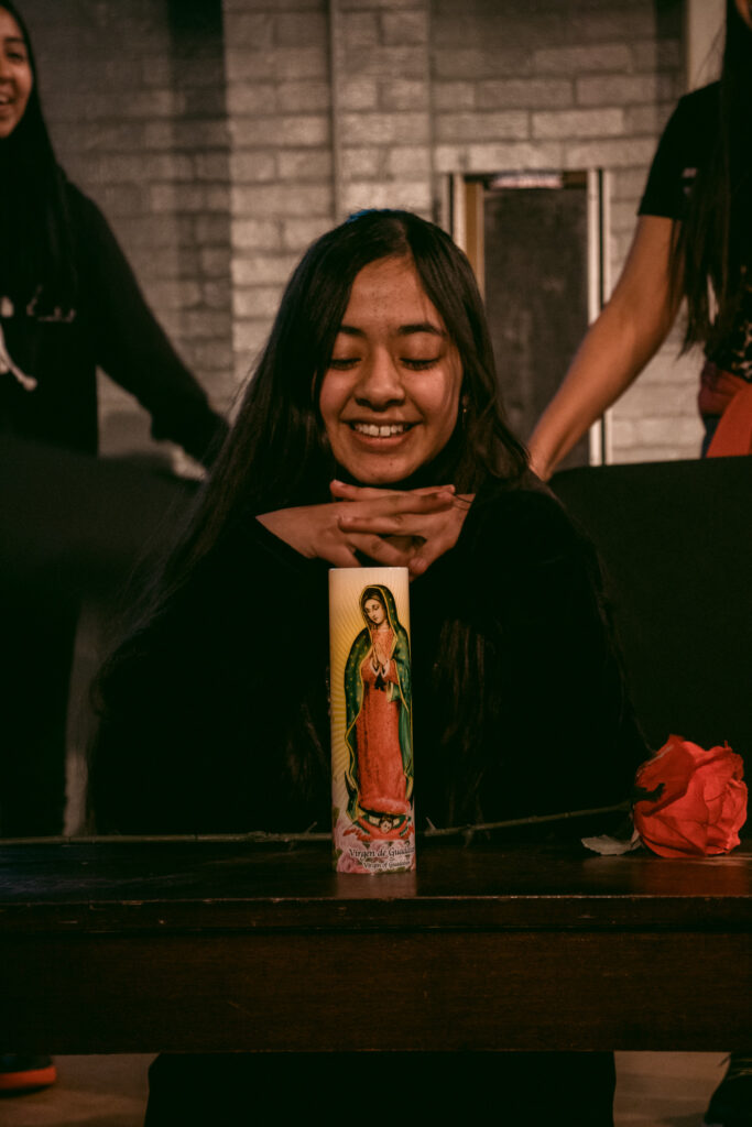 A teenage girl of medium skin tone kneels in front of a candle showing the virgin mary