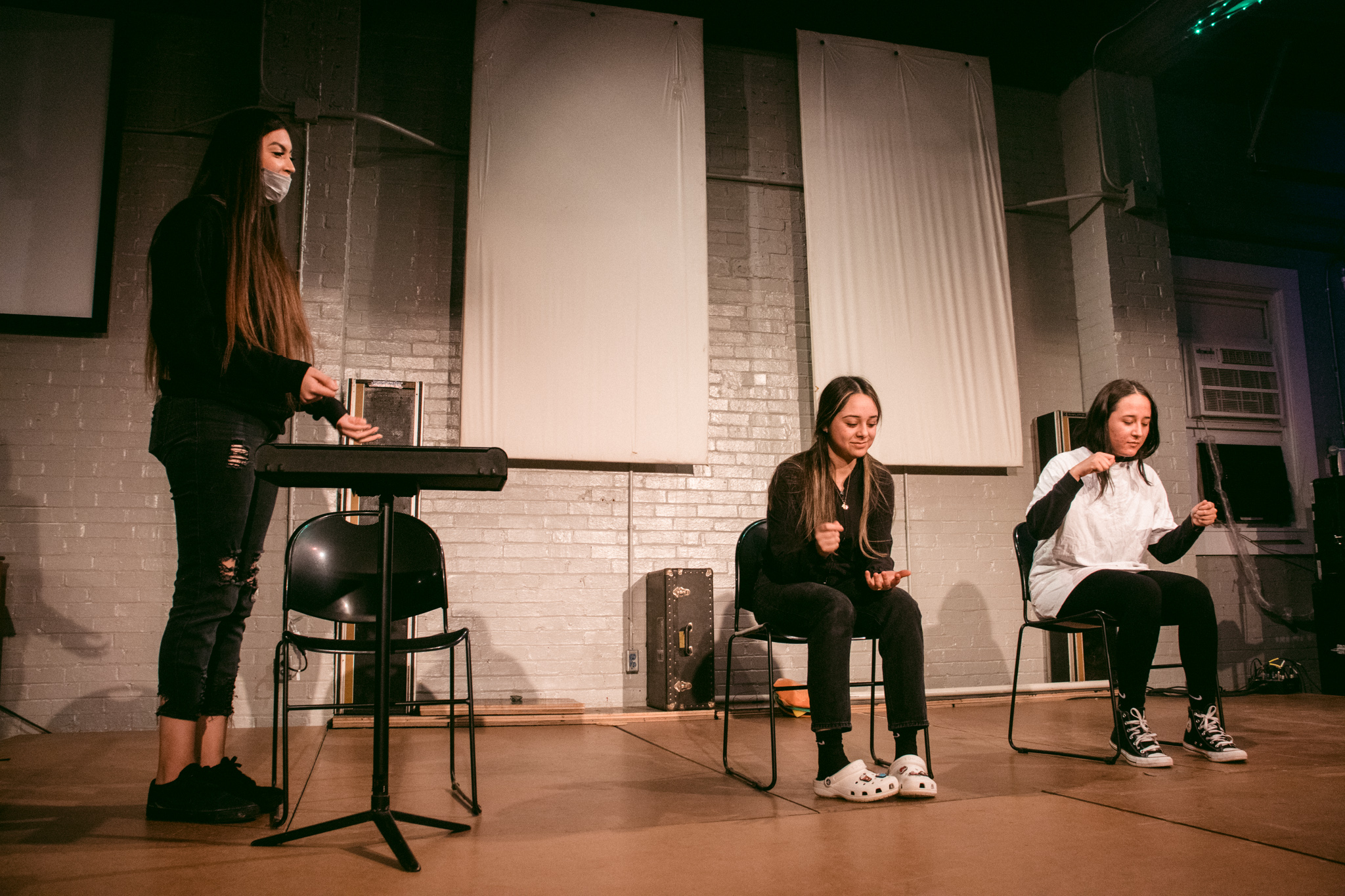 Three teenagers act on a.bare stage sitting on black plastic chairs and one standing at a music stand