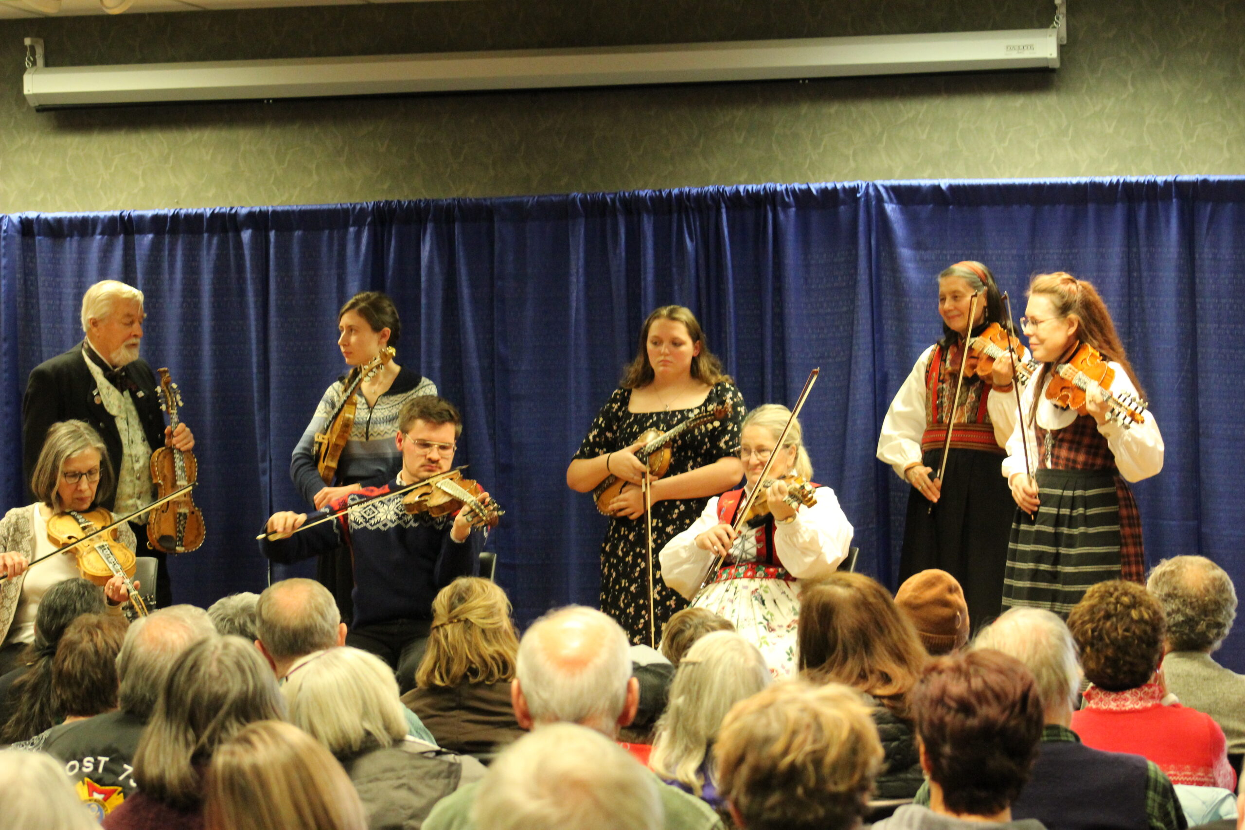 8 people on stage playing intricately decorated fiddles in front of a crowd.
