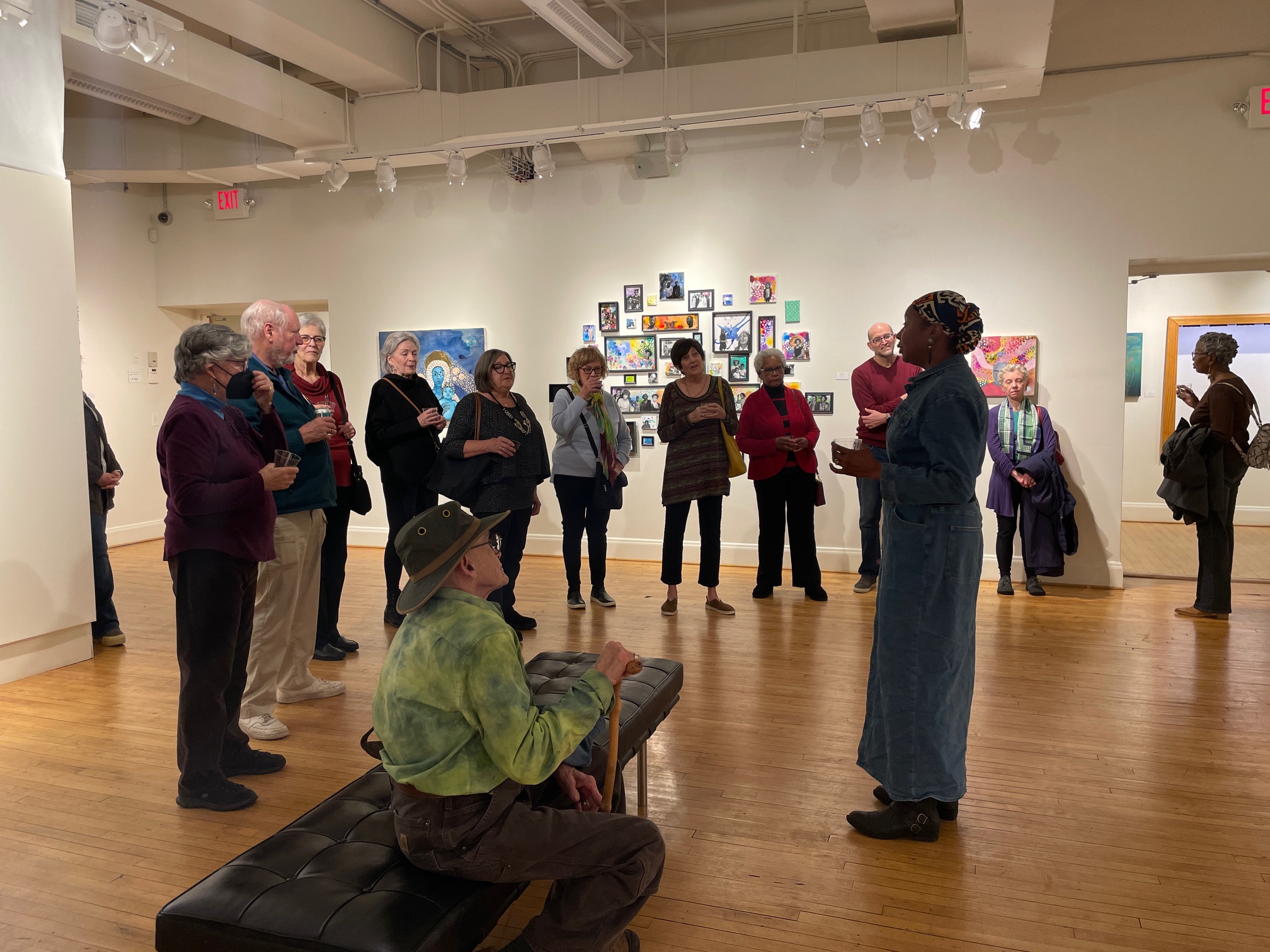 A crowd of people in an art gallery standing around a person of dark skin giving a talk.