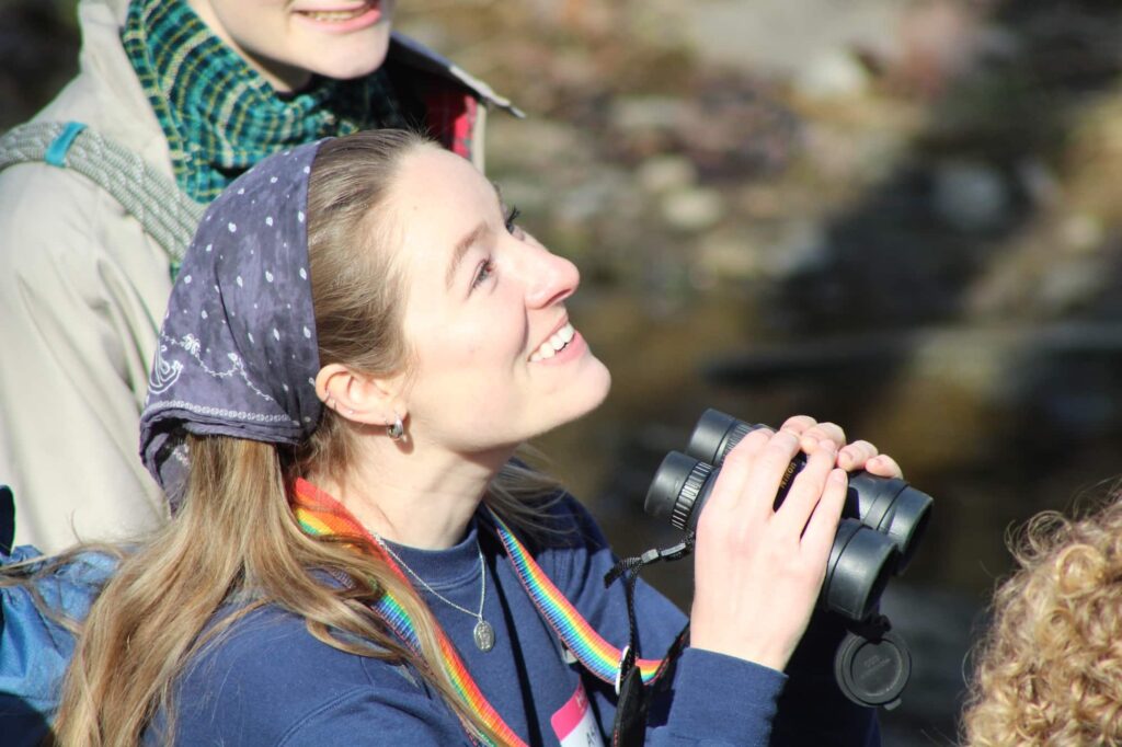 A person with long hair wearing a bandanna on their looks up, holding binoculars at chin-height. The binocular strap has a rainbow pattern.