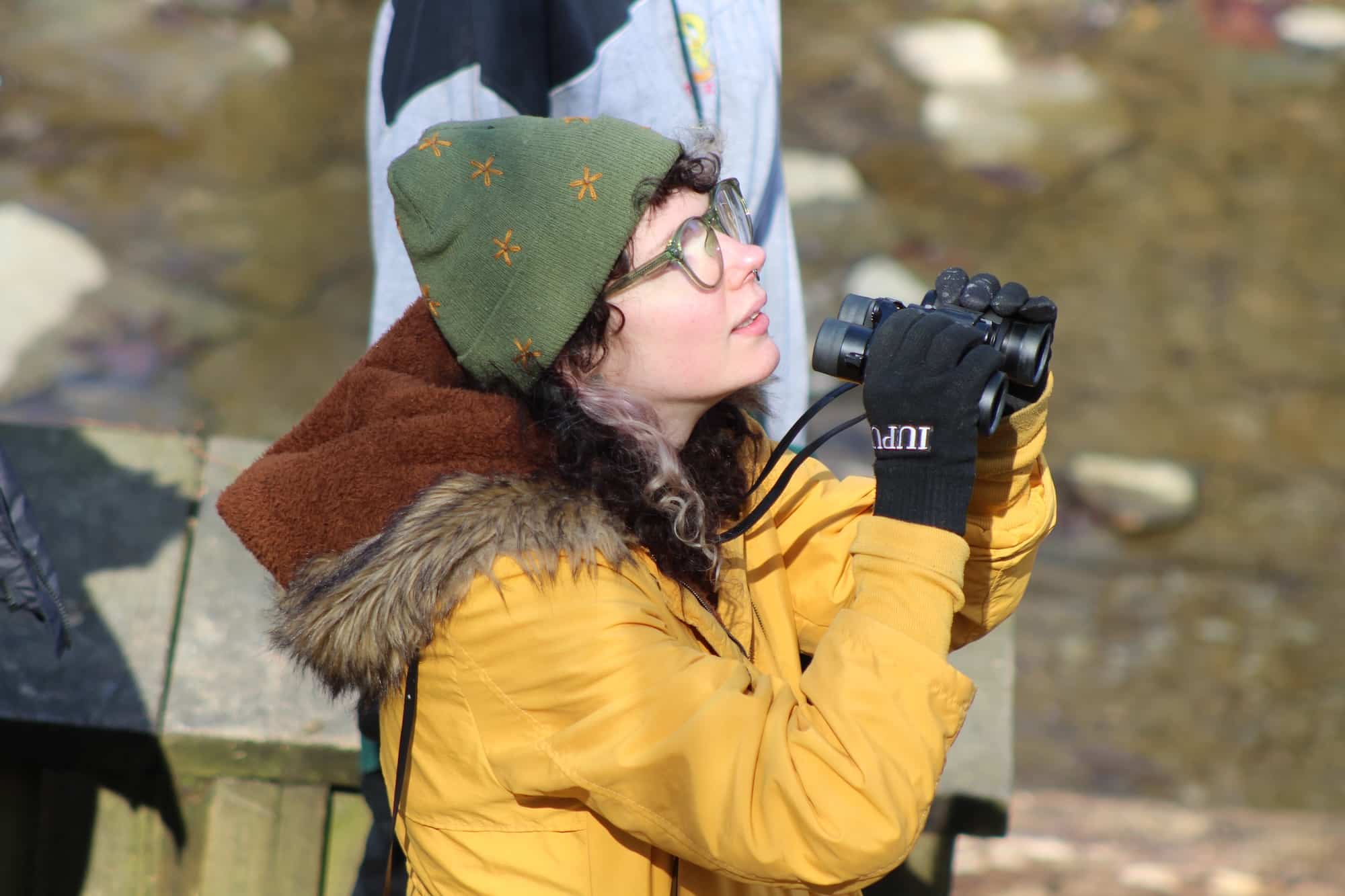 A person wearing glasses and a green hat holds their binoculars down as they look to the sky.