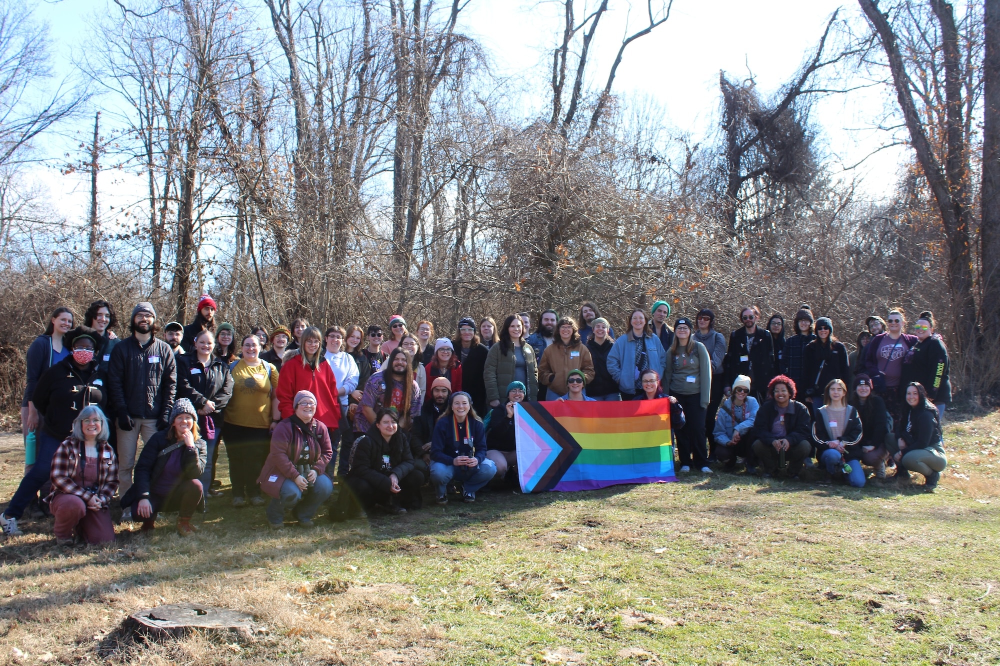 A group of about two dozen people stands smiling in front of a grove of a few barren trees. The people in the front row hold a LGBTQ+ Pride Flag.