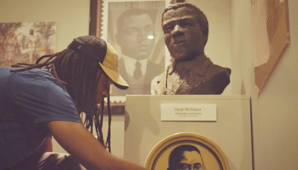 A person with dreads wearing a blue t-shirt and blue and yellow baseball hat bends forward to inspect something in front of a bust on a pedestal.
