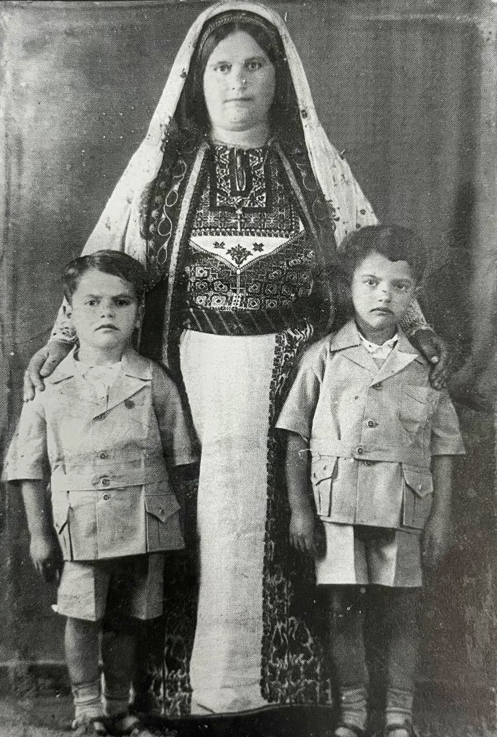 A black and white photo of a person dressed in traditional Palestinian embroidered garment with fabric over her head. She has two young children standing next to her.