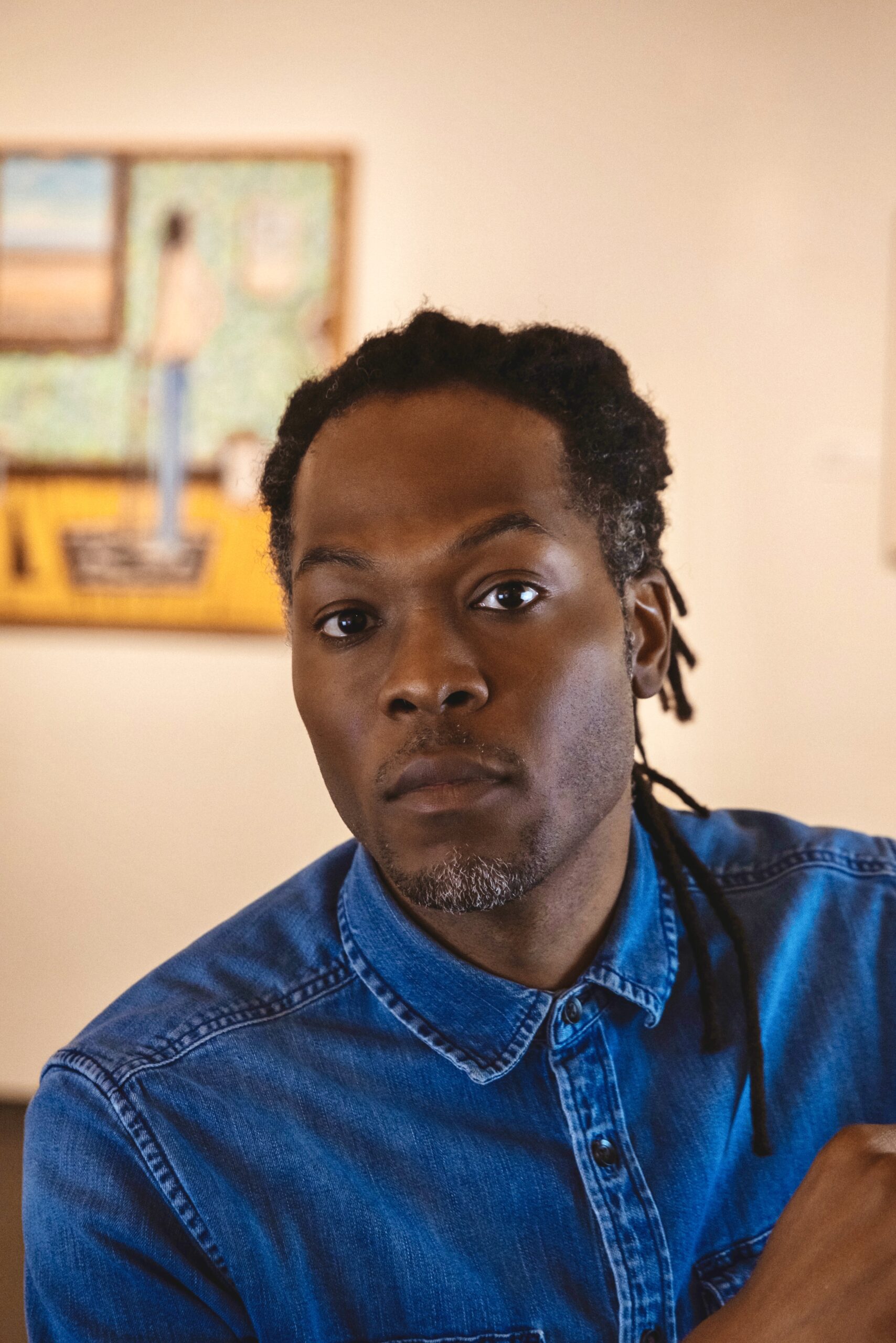 A person in a button up blue collared shirt looks directly into the camera with eyebrows raised. They have their hair pulled back.